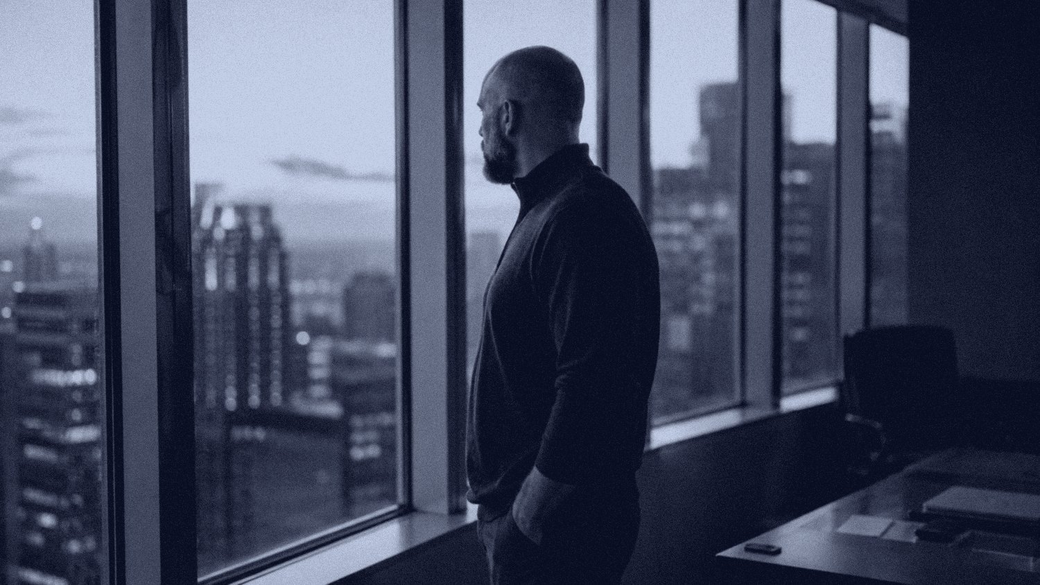 A senior leader alone at a high-floor office window at dusk, weighing a decision